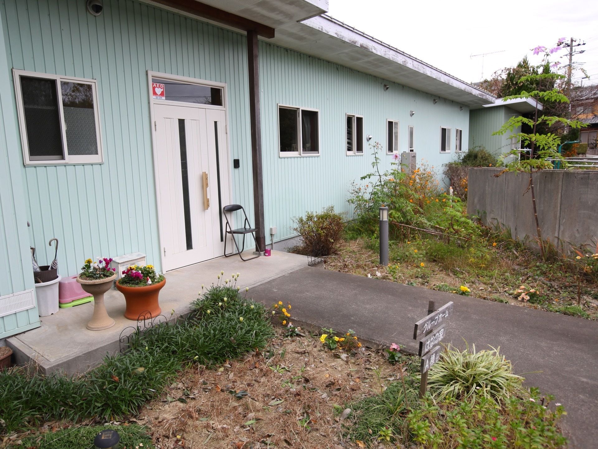 施設入口には、白い片開きの扉が設置され、そばには一脚の椅子やポスト、寄植えされた植木鉢などが置かれている。