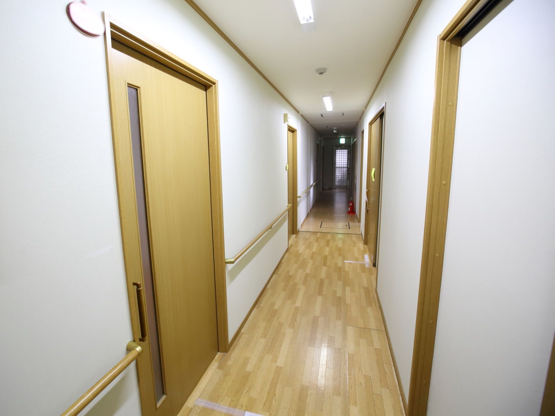 居室前の直線の廊下である。白い壁には手すりが設置され、壁際には防災対策としての消火器が配置されている。
