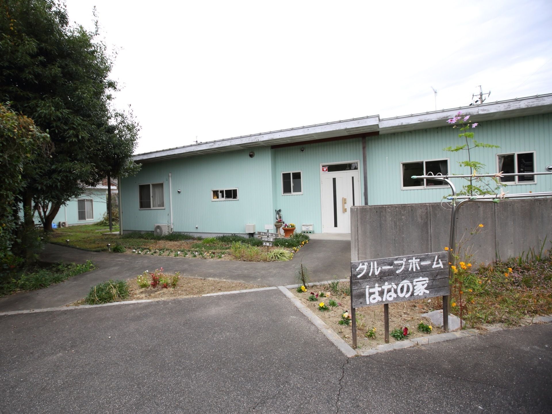 平屋建ての施設外観である。地植えスペースが設けられ草花が植えられている。木製の施設看板のそばに物干し竿が置かれている。