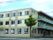 三階建ての学校のような大規模な建物。窓が並んでいる。右手にはベランダがある。角に一本の木が立っている。