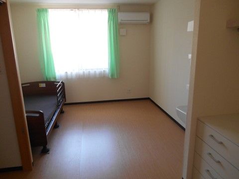 居室には、カーテン付きの広い窓があり、外からの光が差し込んでいる。室内にはエアコン、ベッド、洗面台、タンスが設置されている。