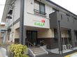 施設の建物の外観である。１階の玄関には４段の階段のほか、手すり付きのスロープがつながっており、植木が植えられている。