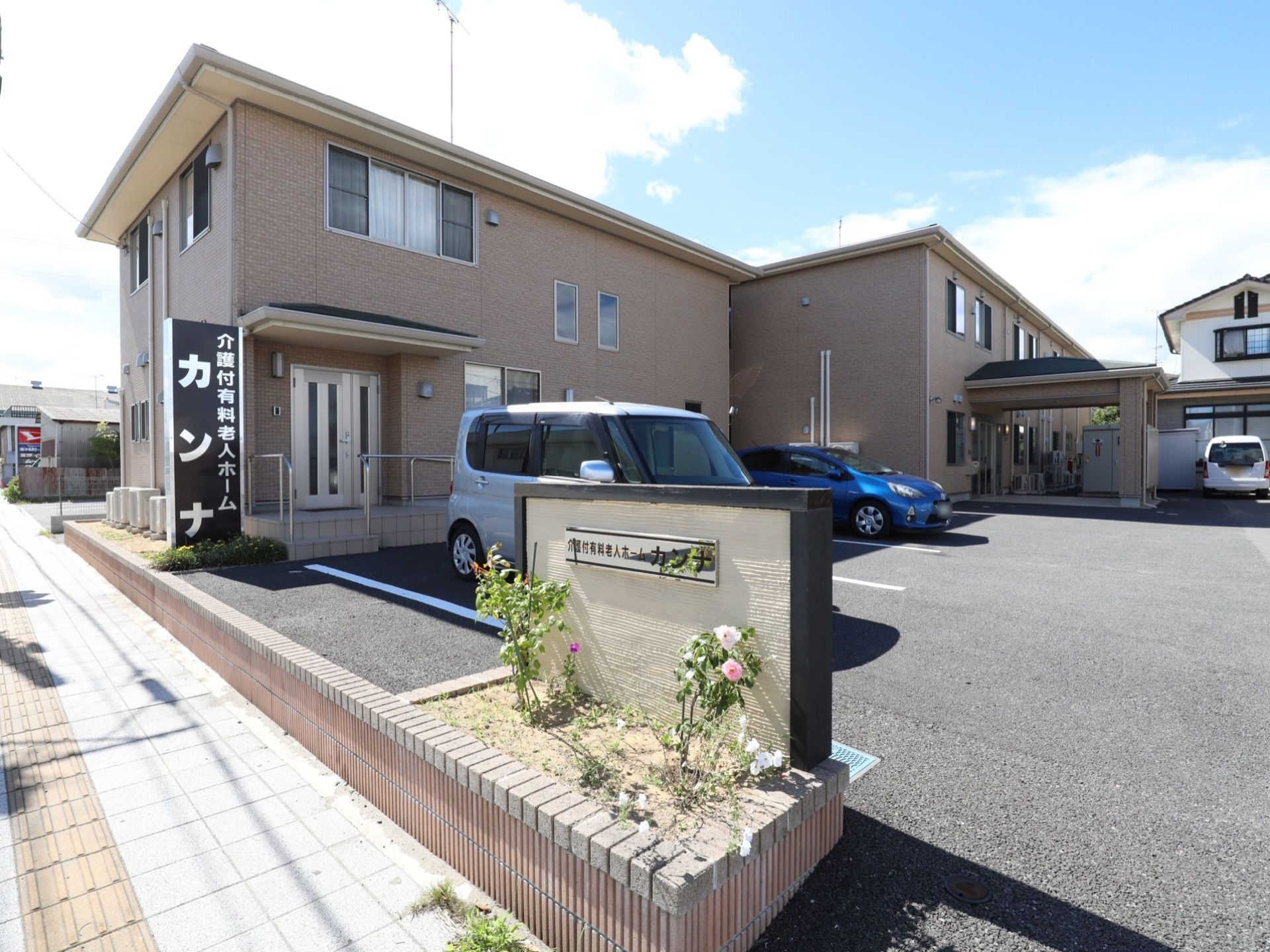 施設の看板の前に花壇があり、花が植えられている。敷地内に車を複数停めることのできる駐車場が完備されている。
