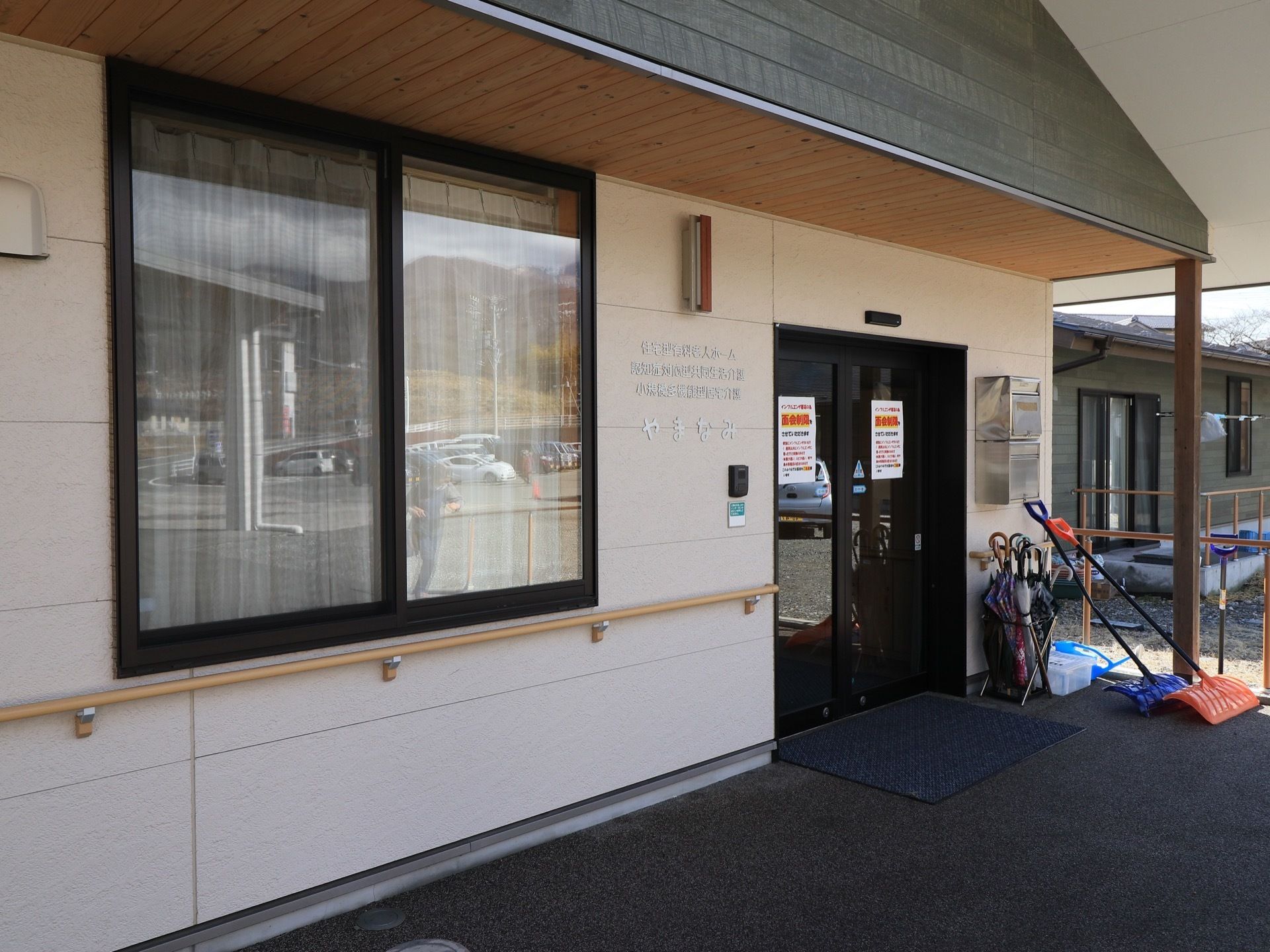 玄関には大きな屋根がついている。黒色の枠のガラス窓の自動扉になっている。玄関前には雪かきの道具が２つ置かれている。