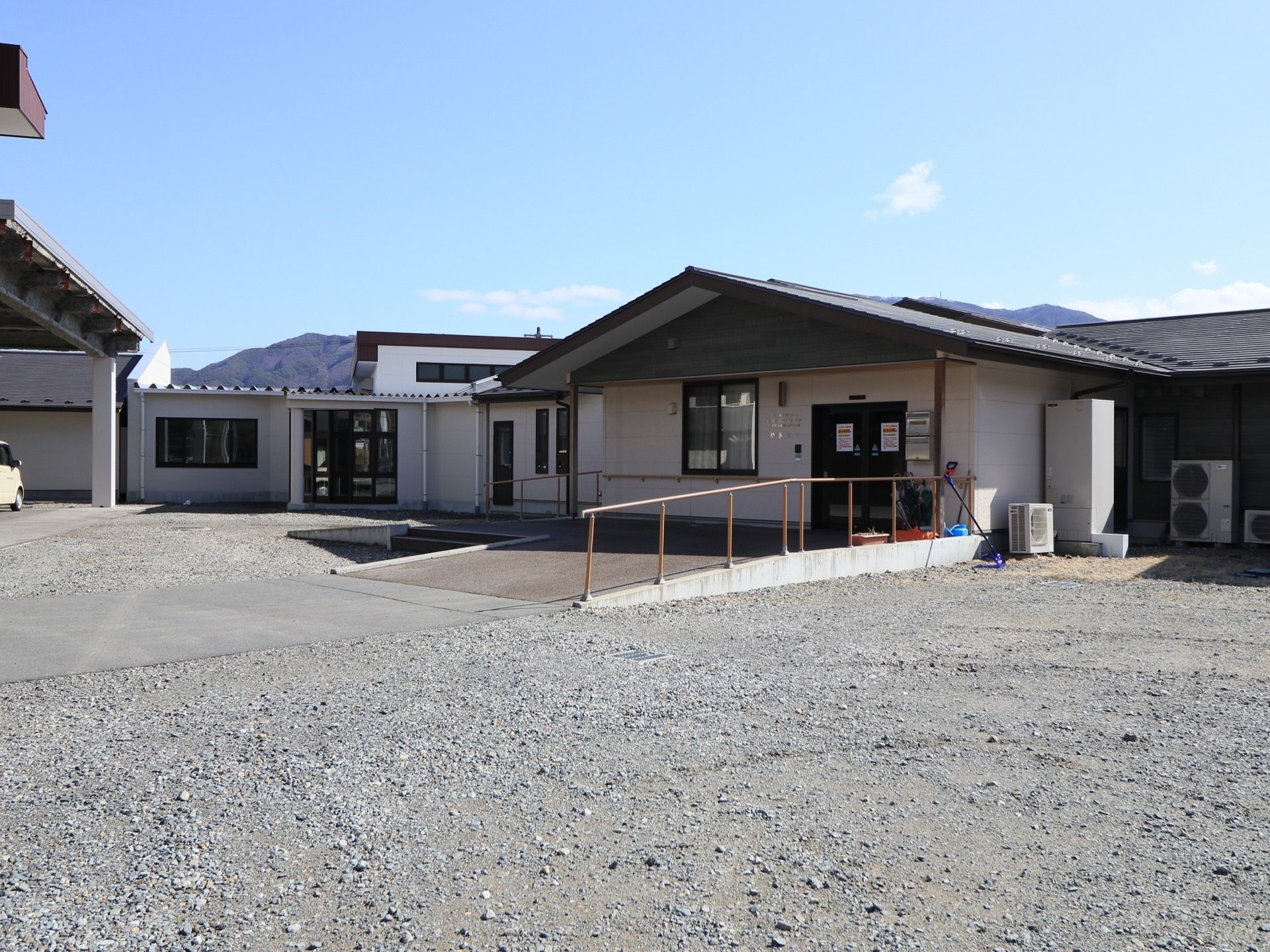 近くに山並みが見えるのどかな環境に立地している施設である。玄関前は幅の広いスロープになっていて、右側には手すりが設置されている。