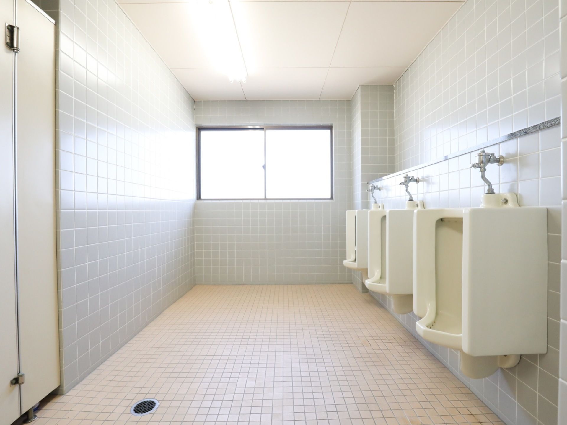 窓から光が射し込む広いトイレは、壁も床もタイル張りである。白い陶器の男性用便器が３据並び、反対側には個室トイレがある。