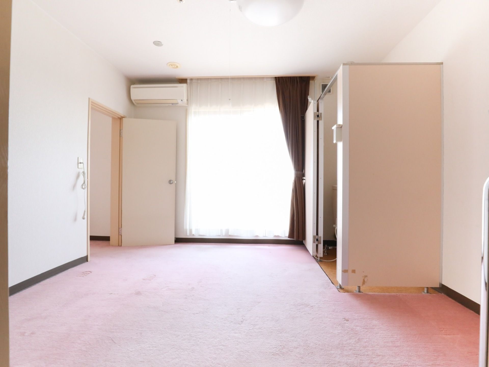 白い壁と床の部屋は大きな地窓から射し込む光で明るい。エアコンとトイレが設置されていて、入口脇には呼び出しボタンがコンセントから下がっている。