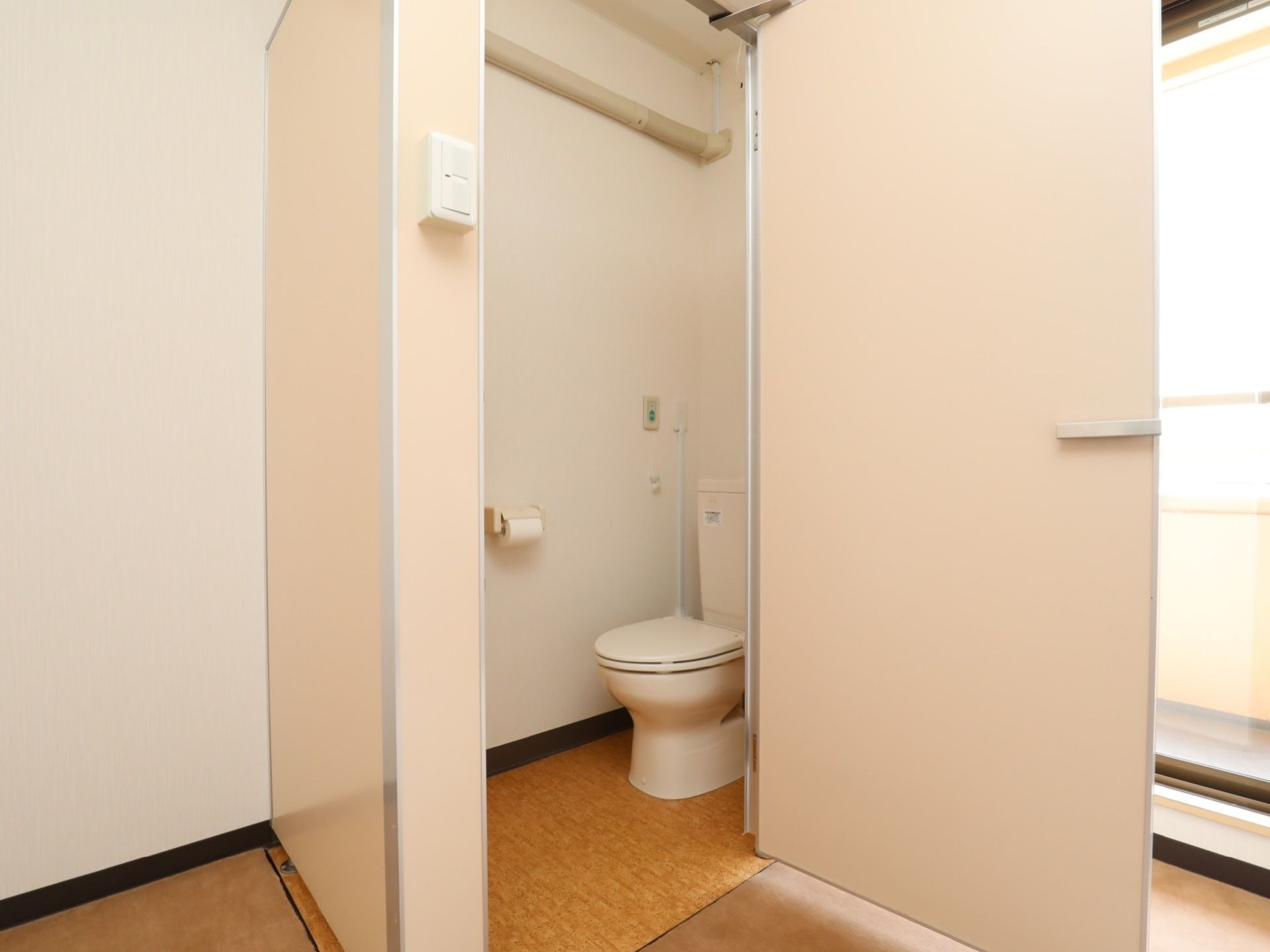 白い部屋の隅に淡いピンクの個室トイレがある。白い洋式便器の脇には、ペーパーホルダーとナースコールがある。