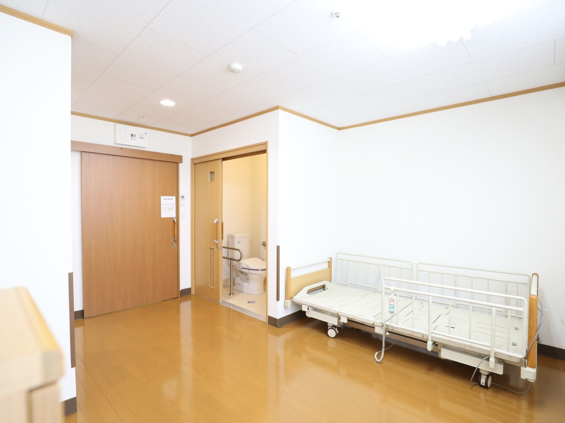 明るく広々とした居室。壁際に電動式ベッドが置かれている。入り口の扉は広く開閉する引き戸になっている。入り口脇にトイレが設けられている。