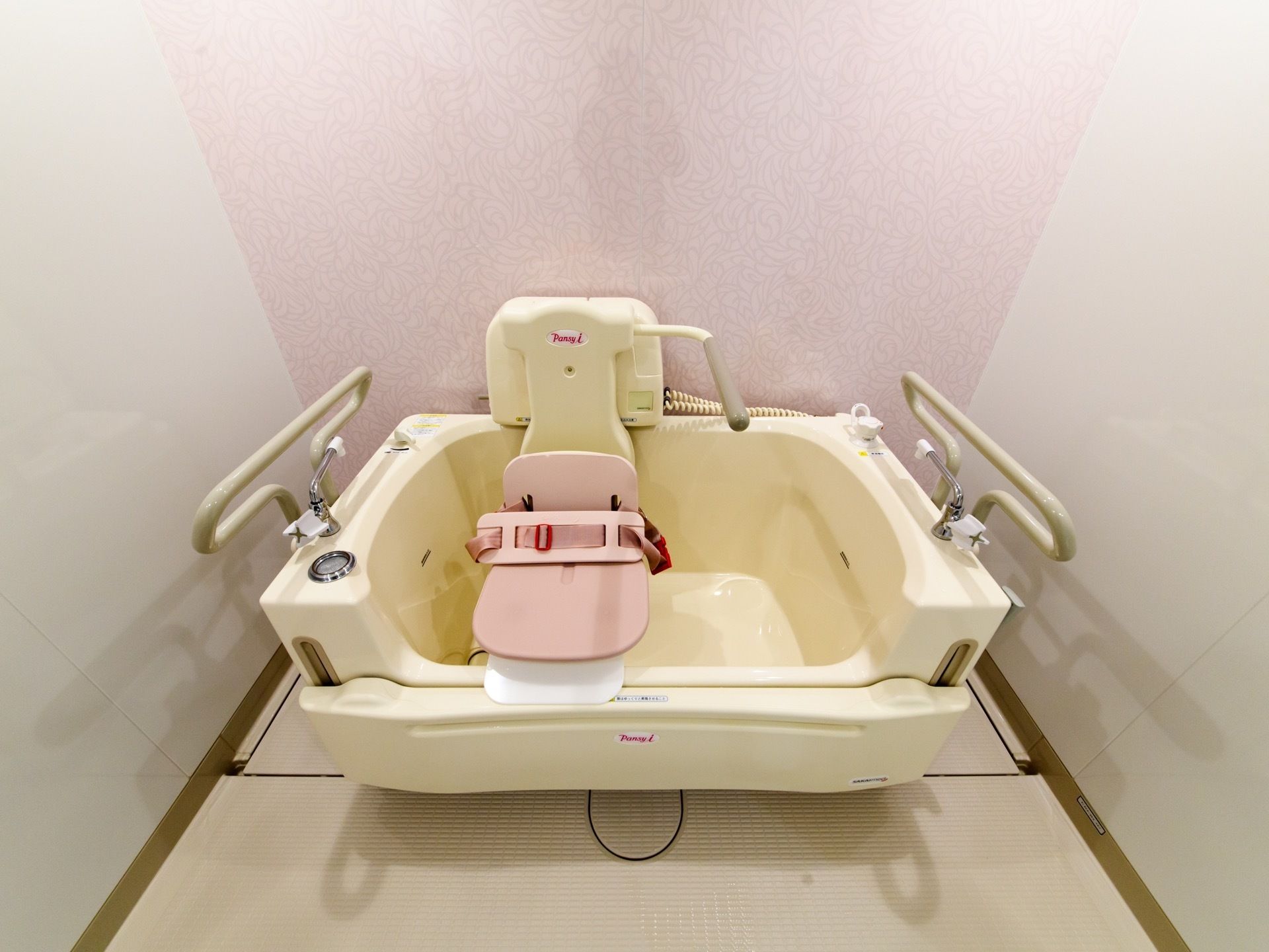機械浴室の壁は、正面の部分だけ淡いピンク色のデザインとなっている。クリーム色のバスタブに、座浴用の器具が付いている。