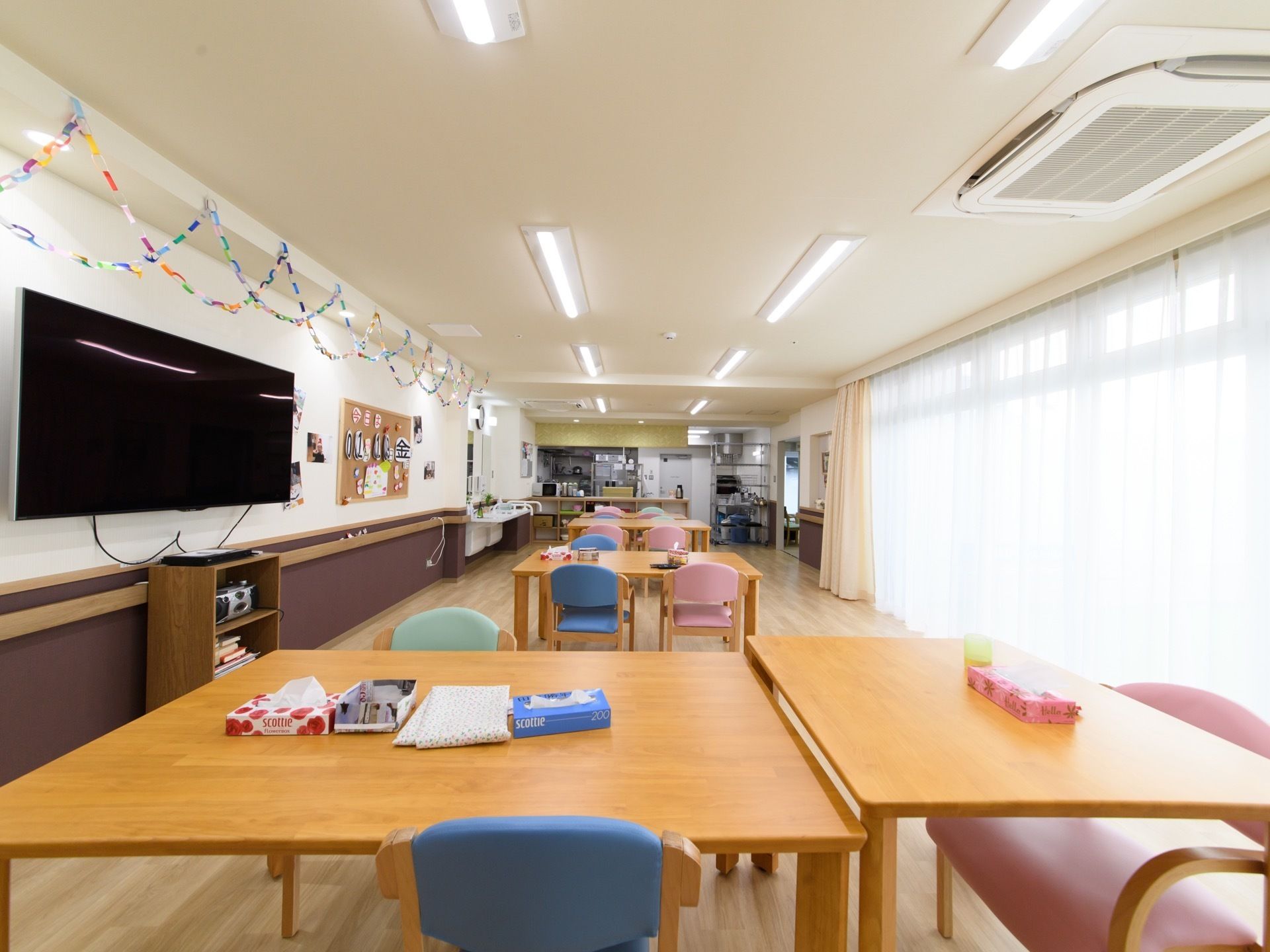 窓から太陽の光が差し込む明るい食堂である。左側の壁には、大型テレビが設置されている。その上は、輪飾りでデコレーションされている。