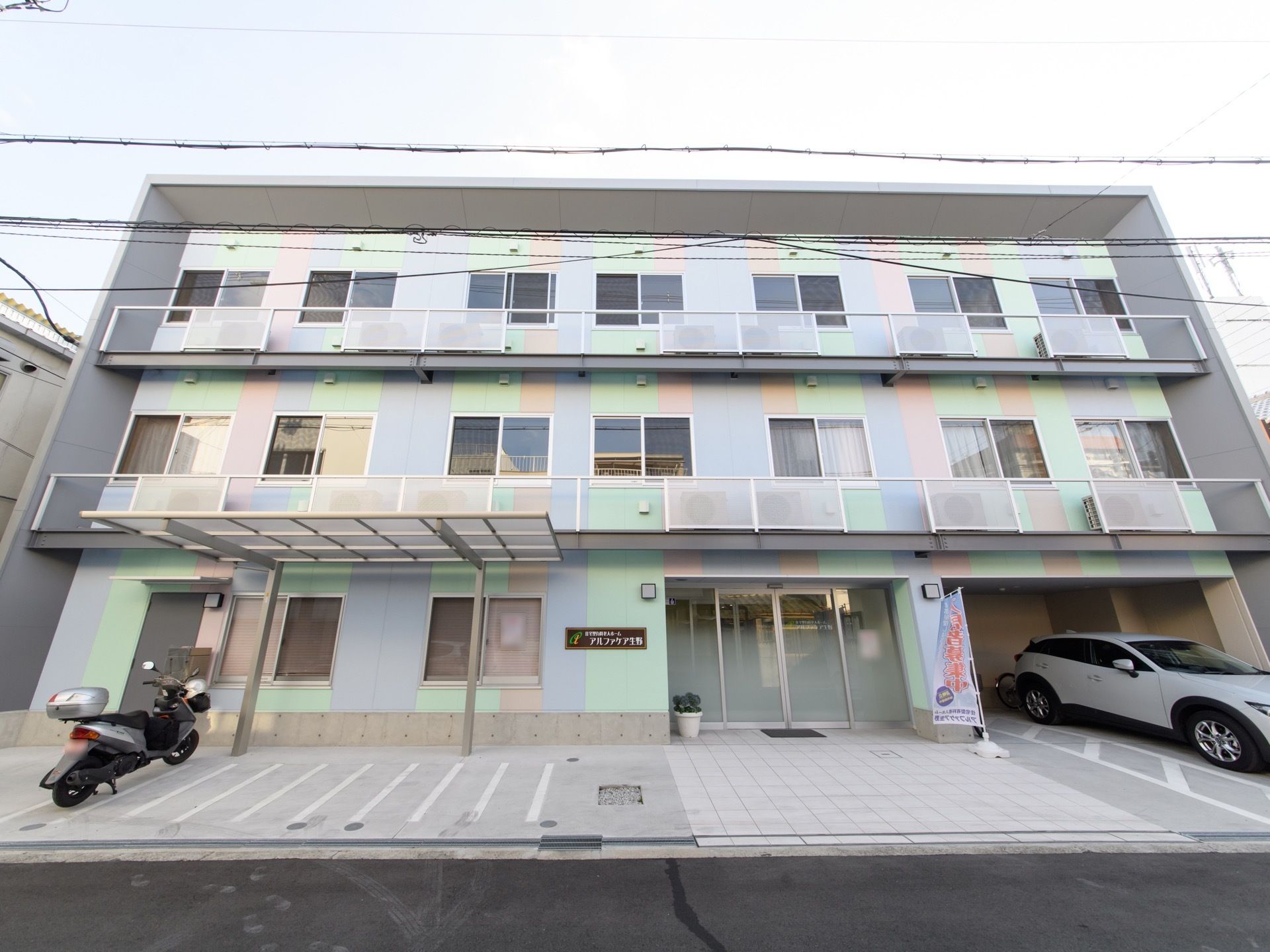 地上３階建ての施設全体を、正面から写したものである。左端には屋根付きの駐輪場があり、バイクが１台置いてある。