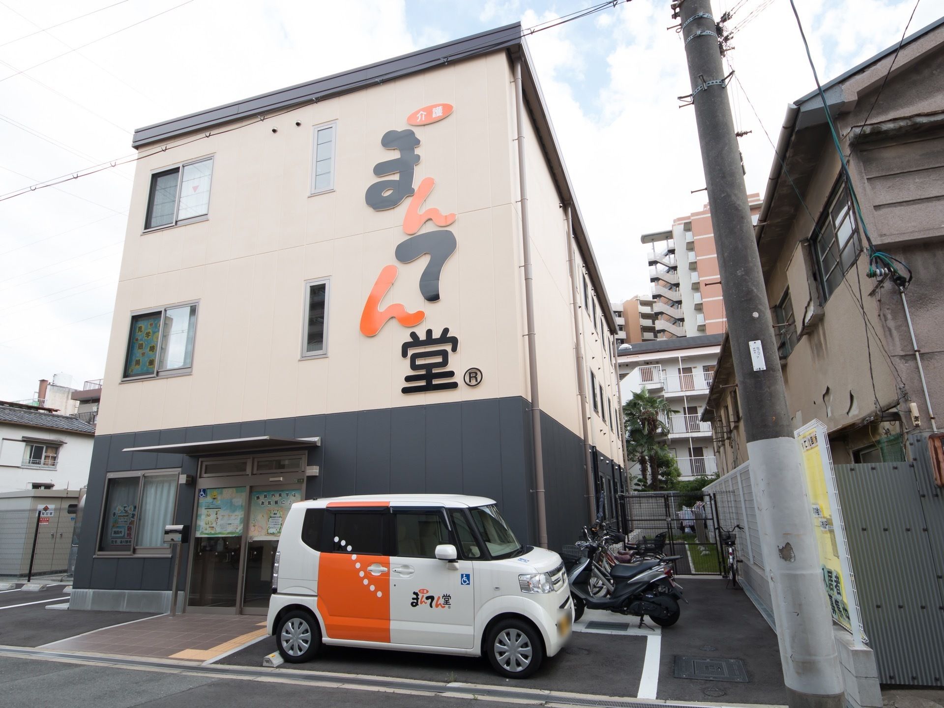 ベージュと黒の外壁で、通りに面した壁には大きくホーム名のロゴが書かれている。エントランス前には駐車スペースがある。
