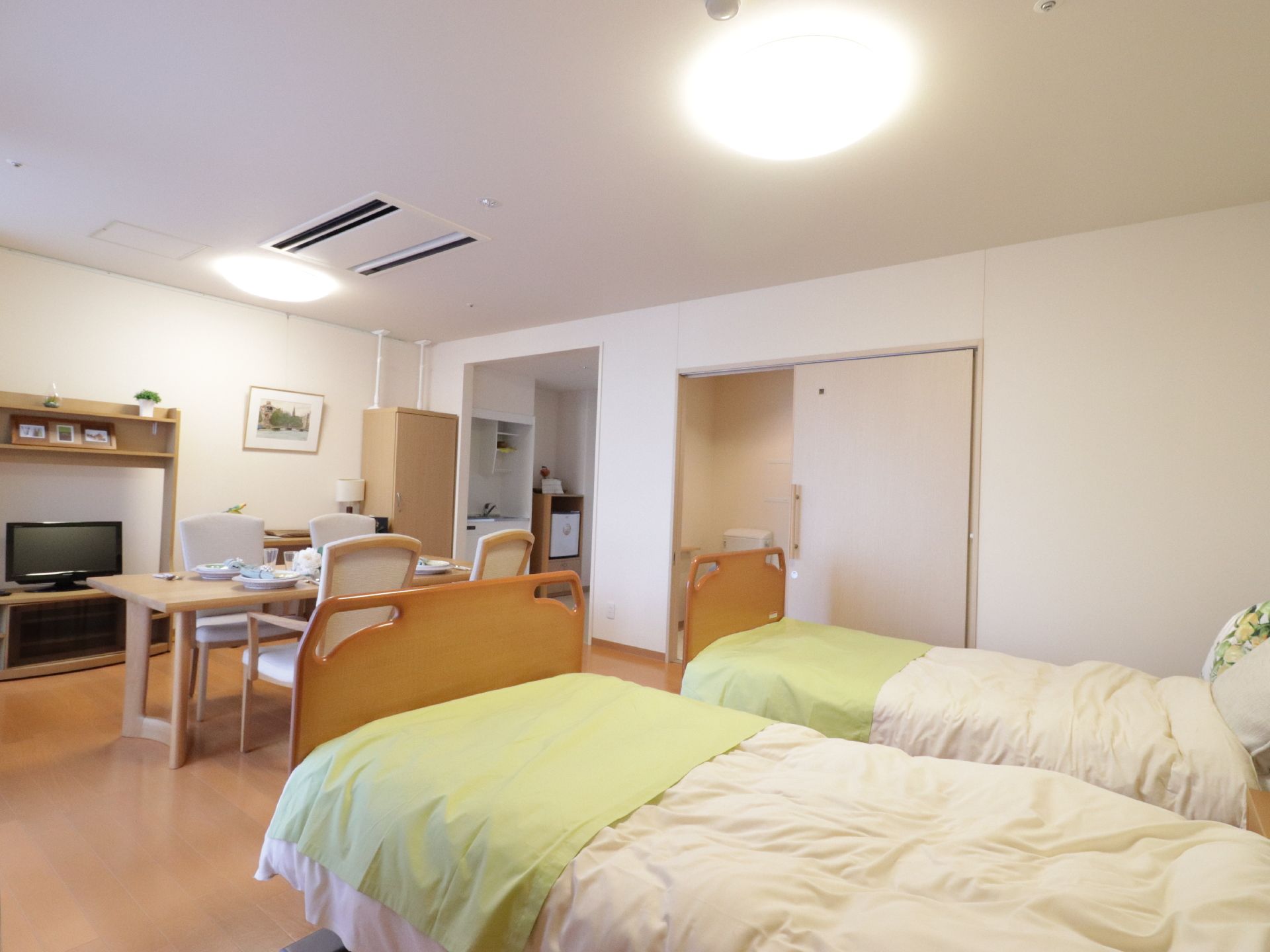 白色と木目調の内装の２人用の居室には、ミニキッチン、トイレ、エアコン、介護用ベッドなど、生活に必要な設備を整えている。