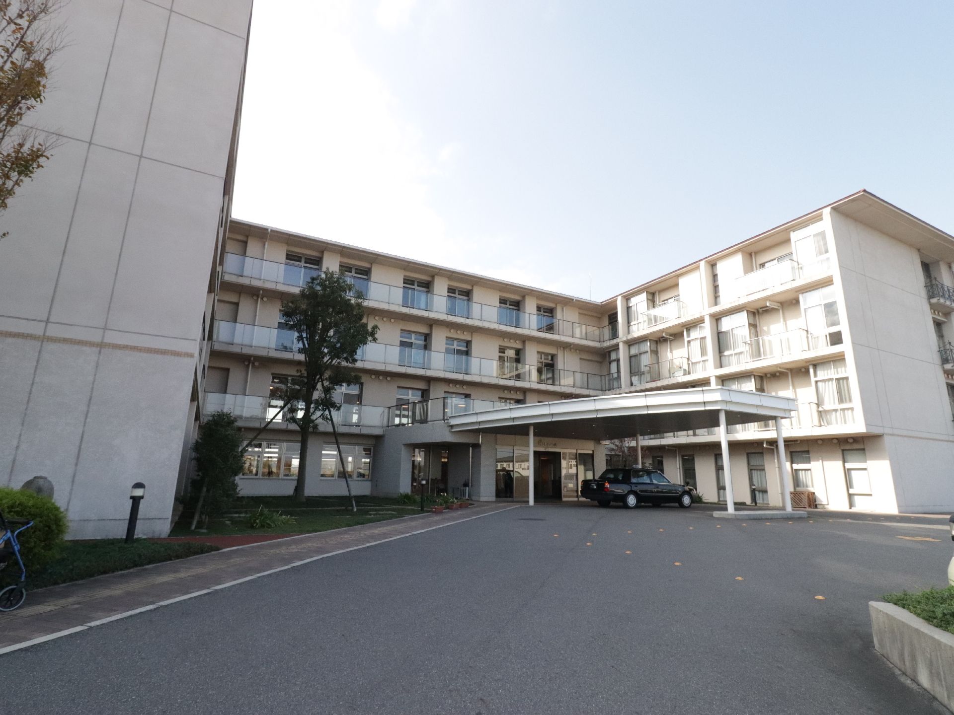 白色の外観の４階建ての建物である。玄関前には、大きな屋根があり、車が１台停まっている。横側には植栽を施している。