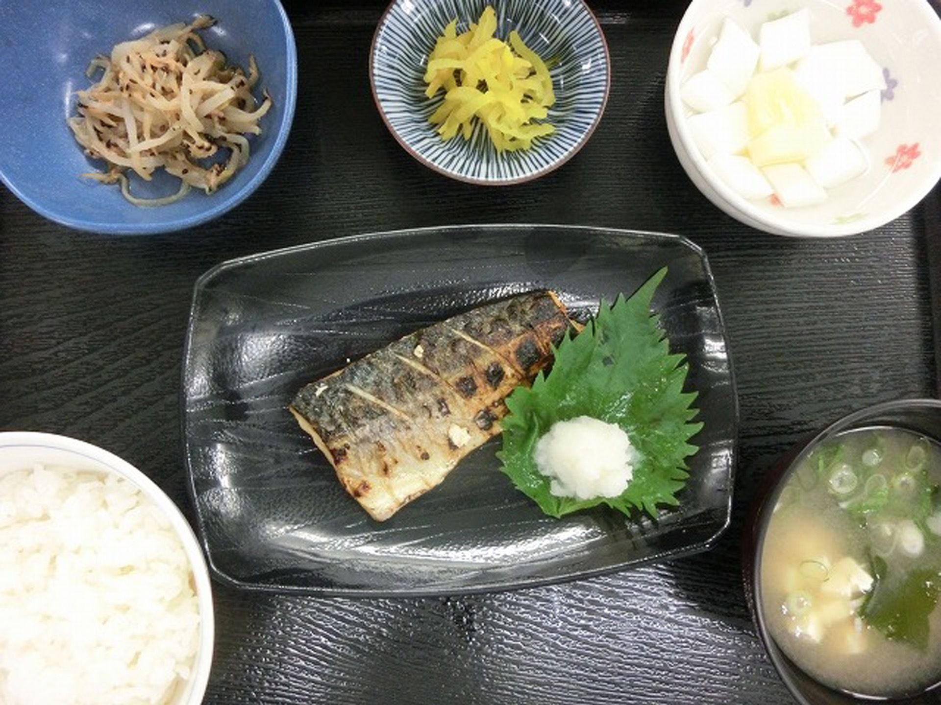 黒色のトレイに、メインの焼き魚を中央に置き、ご飯とお味噌汁、小鉢を３品置いて並べている。品数豊富な食事である。