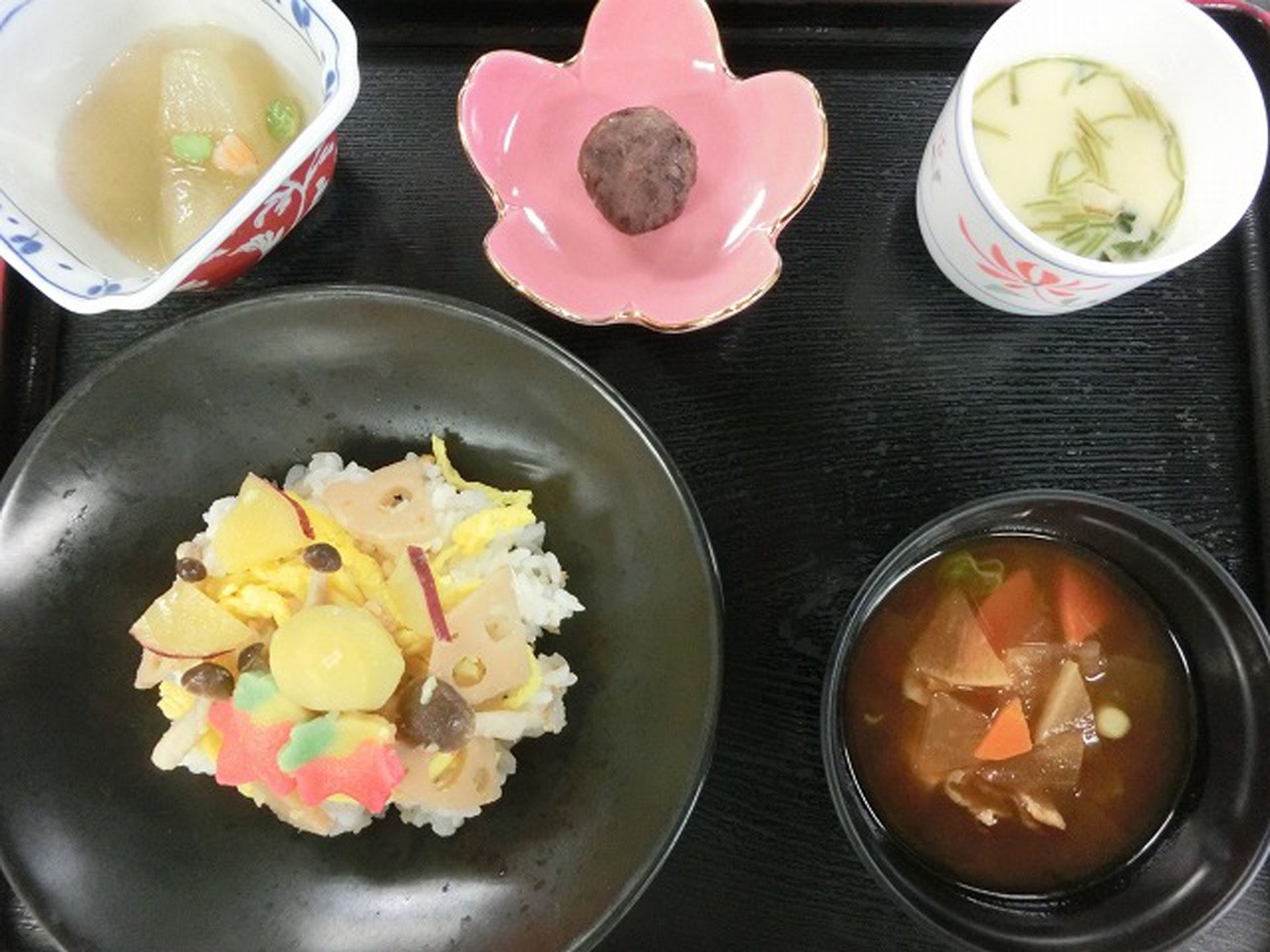 黒色のトレイの上に混ぜご飯に汁物、３品の小鉢を添えている。食器にも配慮した、彩りのキレイな食事である。