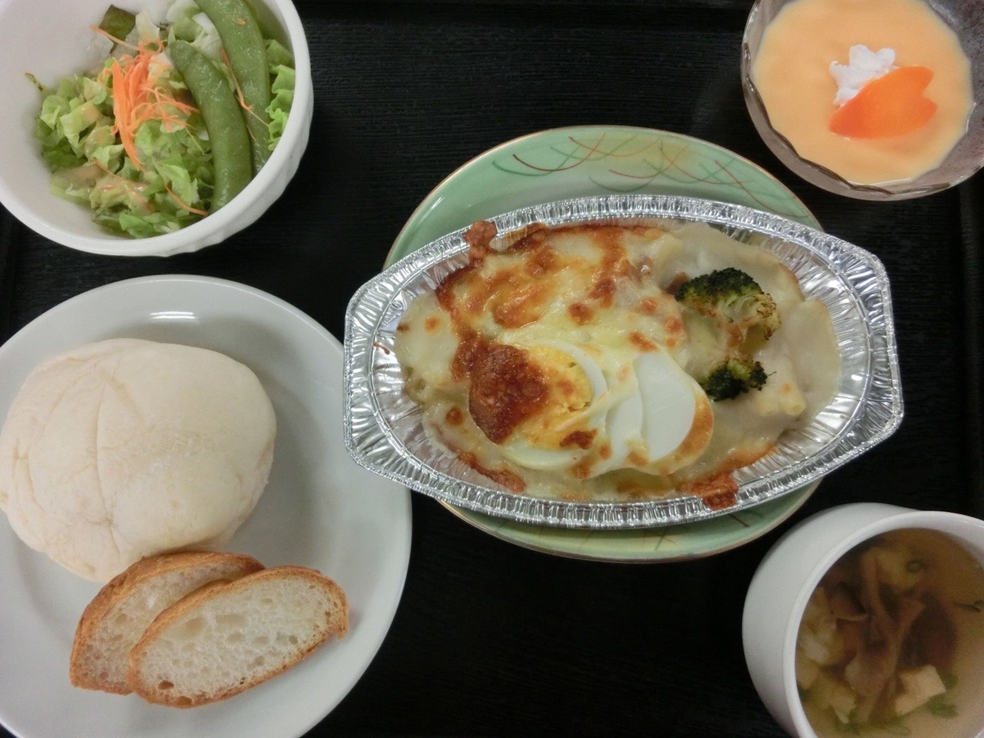 中央にグラタン、手前のお皿には２種類のパン、そのほかにスープと野菜、デザートを添えたパン食の献立である。