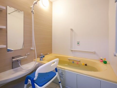 ユニットバスタイプの浴室の洗い場に青いシャワー椅子が置かれている。黄色い浴槽の中に４つのおもちゃが浮いている。