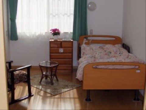 居室には木製のベッドやチェストのほか、椅子とテーブルが置かれている。グリーンのカーテンが取り付けられている窓辺には、花が飾られている。