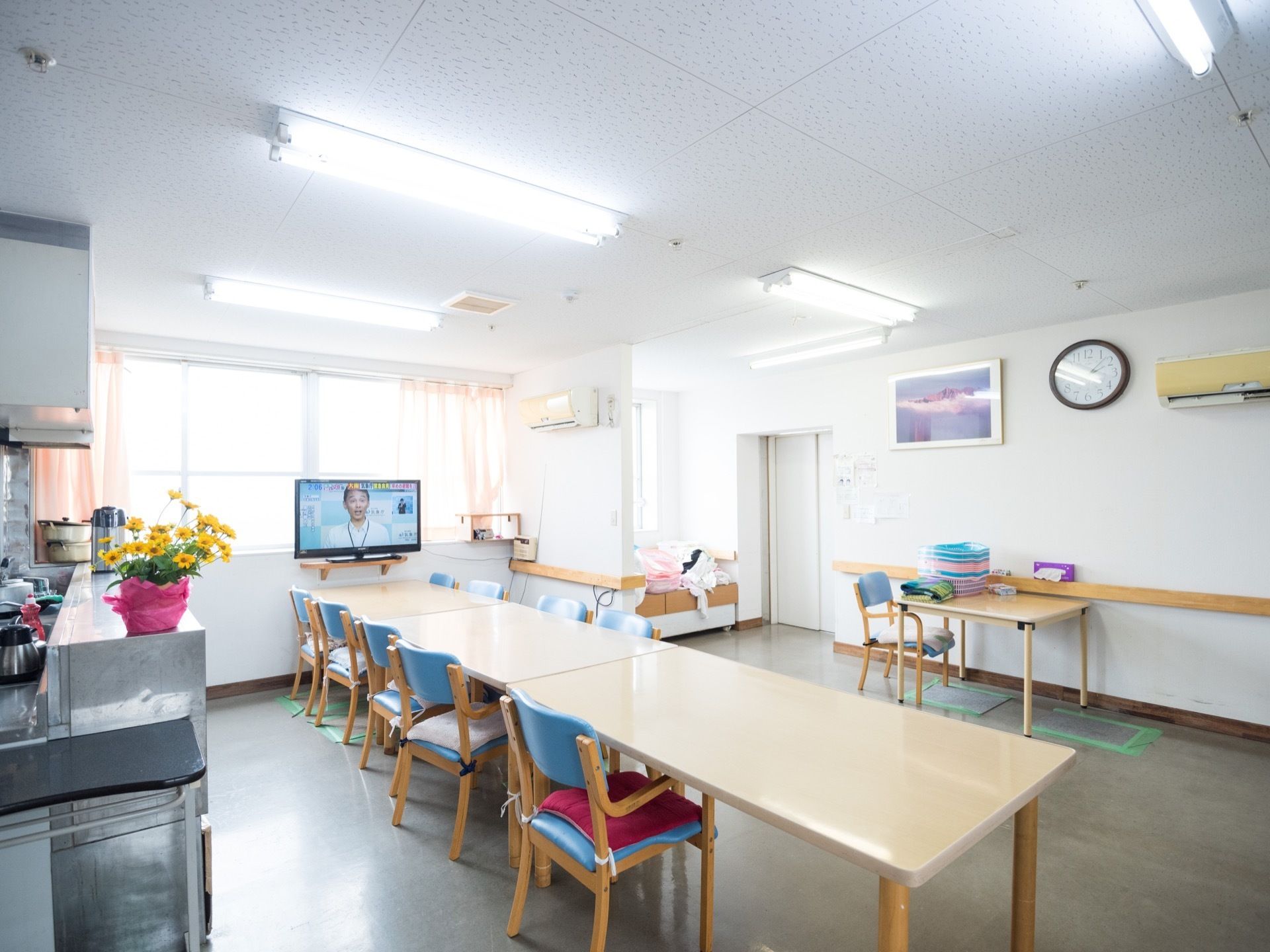 キッチンのすぐ目の前に、テーブルが３つ並べて置いてあり、その側にはテレビが取り付けられている。壁には、絵画やエアコン、時計が設置されている。