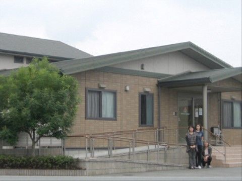 緑色の三角屋根の建物である。玄関には屋根があり、階段かスロープで上がることができる。階段下には、３名の女性スタッフがいる。