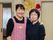 女性スタッフ２名が、肩に手をかけた状態で笑顔で写っている。背景の壁には、色づいた葉を表した作品が飾られている。
