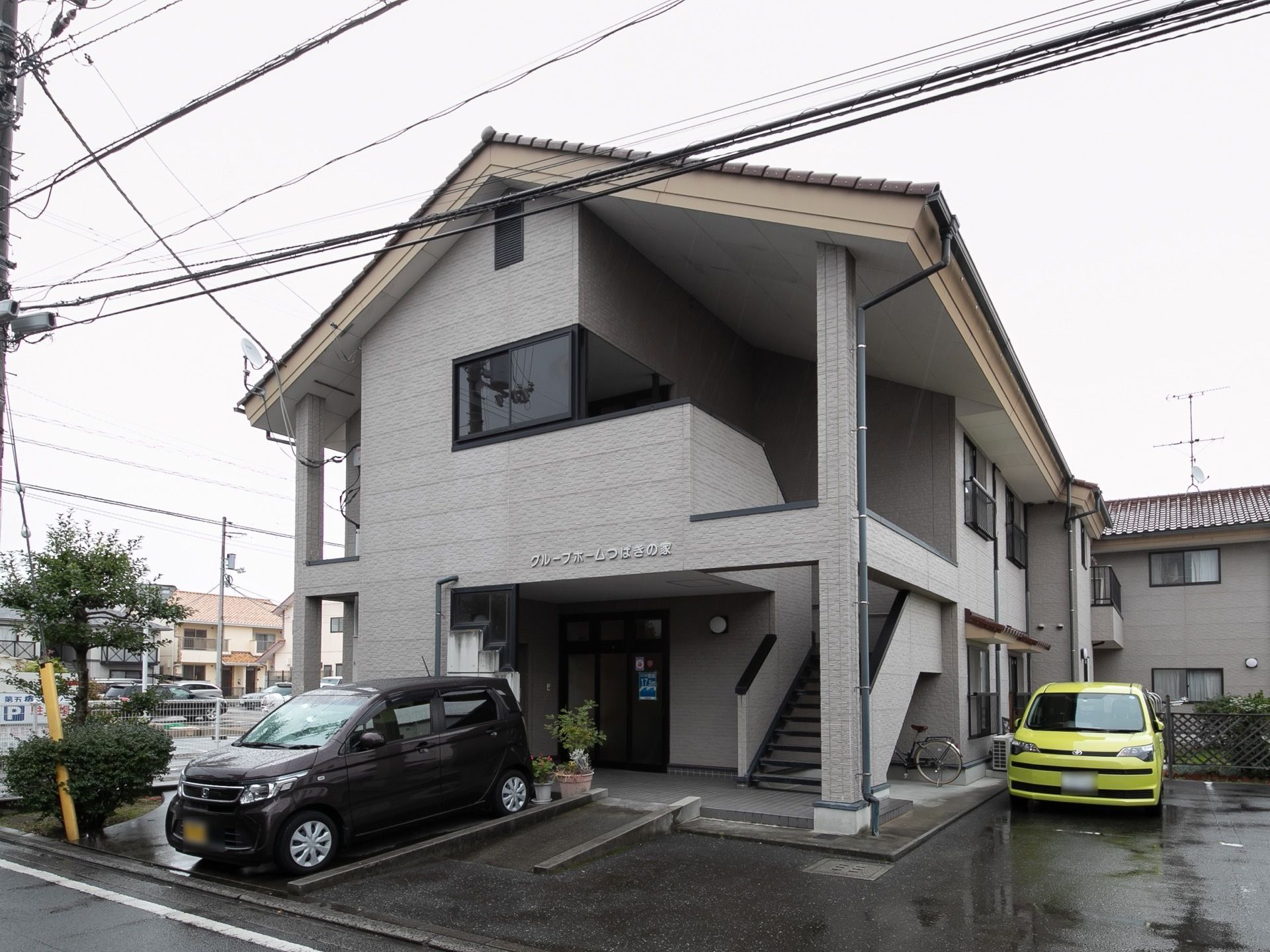 灰色を基調とした外壁の２階建ての施設である。玄関前にゆるやかなスロープがあり、その横に車が停められている。