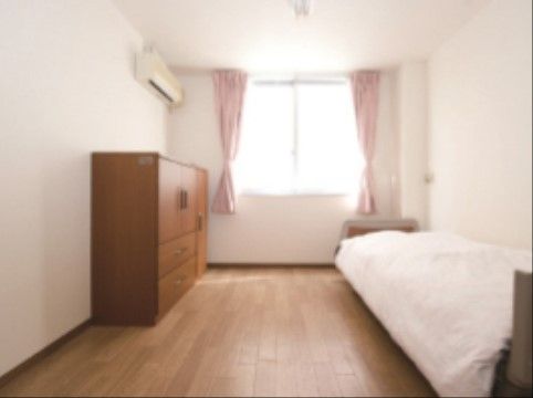 居室の床は木製のフローリングである。奥に窓があり、左側の壁にエアコンが設置されている。ベッドやタンスが置かれている。