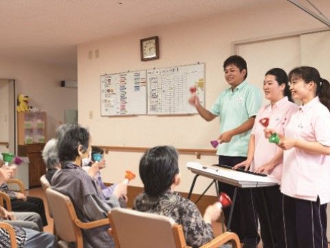 手前に入居者様たちが椅子に座っていて、手にハンドベルを持っている。正面に３人のスタッフが立っていて、ハンドベルを教えている。