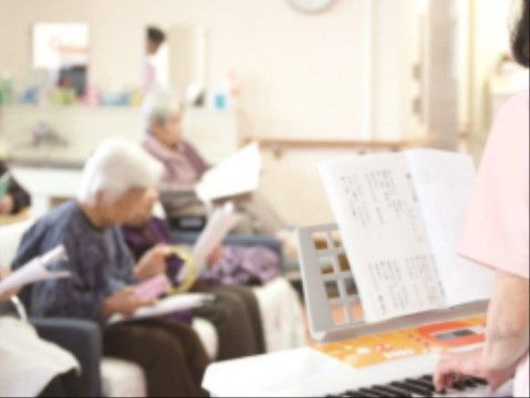手前にキーボードが設置されており、女性が演奏している。その向こう側に入居者様たちが座って、歌詞を見ながら歌っている。