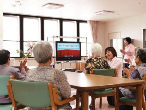 日差しが入る明るい室内で、大画面のテレビに向かい、４名の入居者様が座っておられる。スタッフ２名が付き添っている。