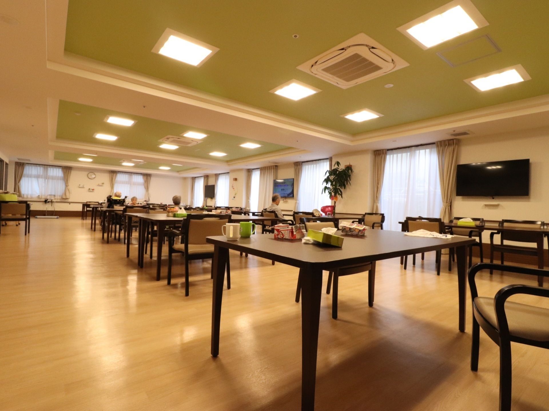 多くの入居者様が集まれる大食堂。大型の壁掛けテレビを窓際に何台も設置している。エアコンは天井に取り付けられている。