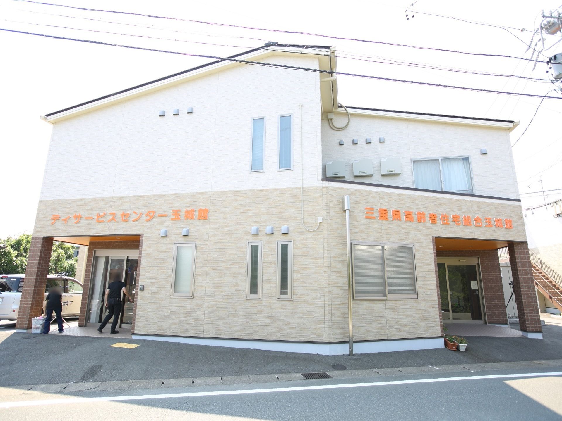 施設はベージュ色の外壁で２階建ての建物である。別に入口が設けられたデイサービスが併設されており、入居者様も利用できる。