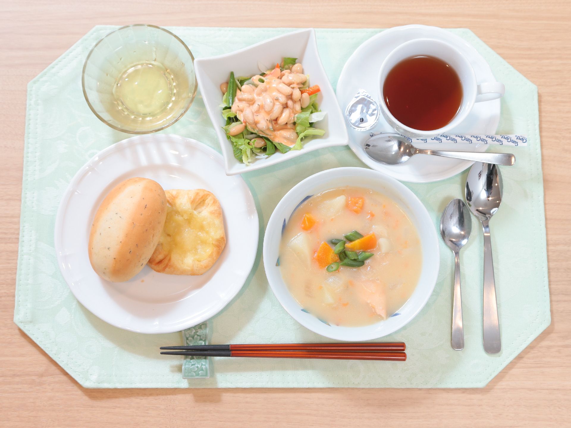 野菜たっぷりのホワイトシチュー、レタスと大豆のサラダ、丸いロールパン、紅茶などが、白いトレーに並べられている。
