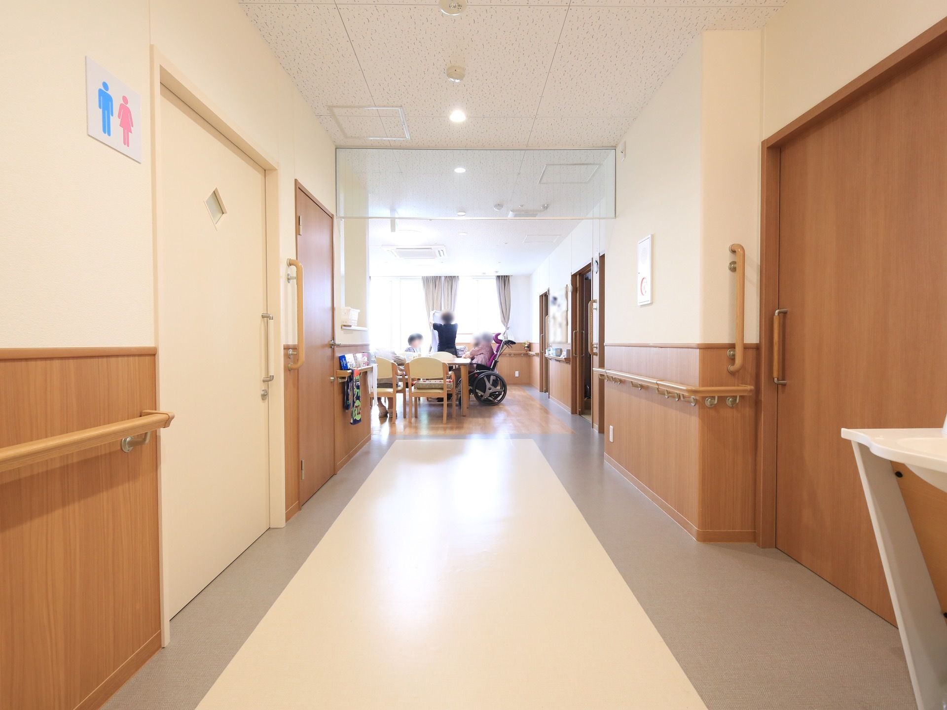トイレ前の直線廊下は、大きなガラス窓があるリビングに繋がっている。リビングでは入居者様がテーブルを囲み集っている様子が、廊下からよく見える。
