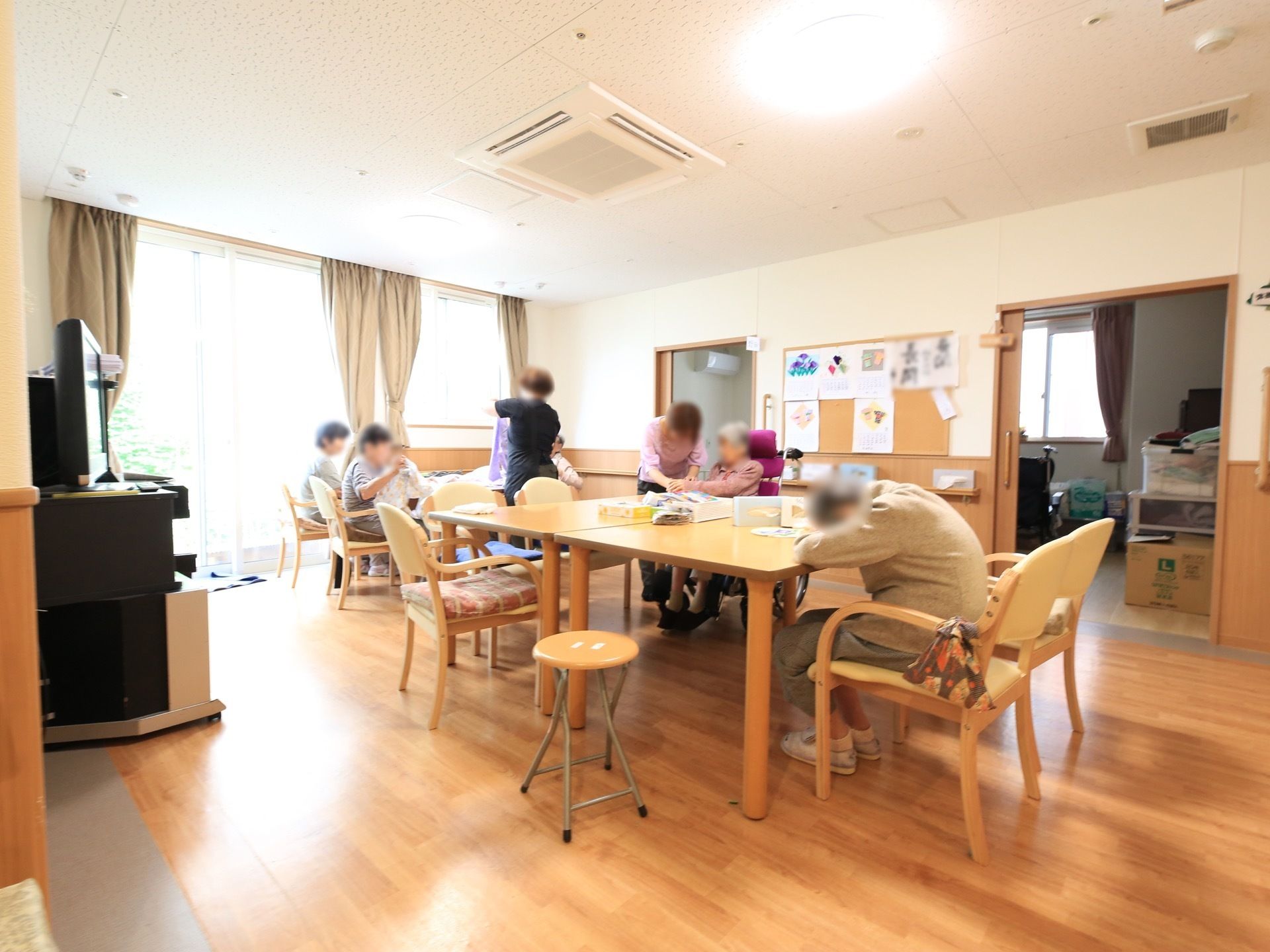 リビングは、一面が天井までガラス張りなので明るい空間である。部屋の中央には、テーブルと椅子が複数配置され、入居者様数人が着席している。