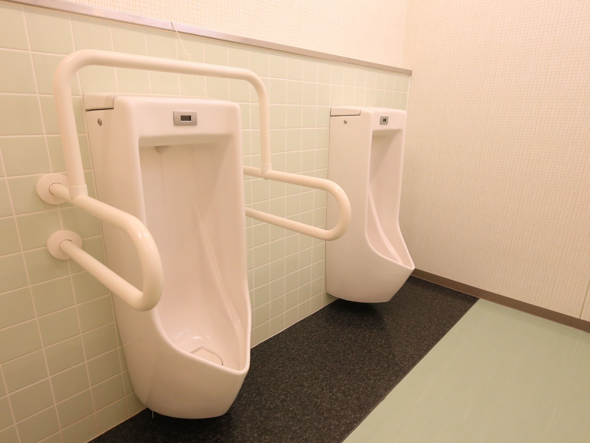 手すりのついた共用の男子用トイレ。壁や床は全体的にタイル張りになっており、小便器の足元だけは黒い御影石製をしている。