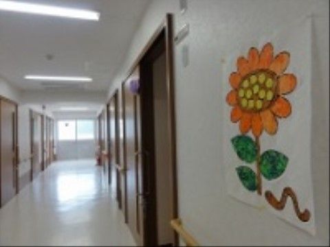 居室が立ち並ぶ明るい廊下である。壁にひまわりの絵が飾られており、両サイドには木製の手すりが設置されている。