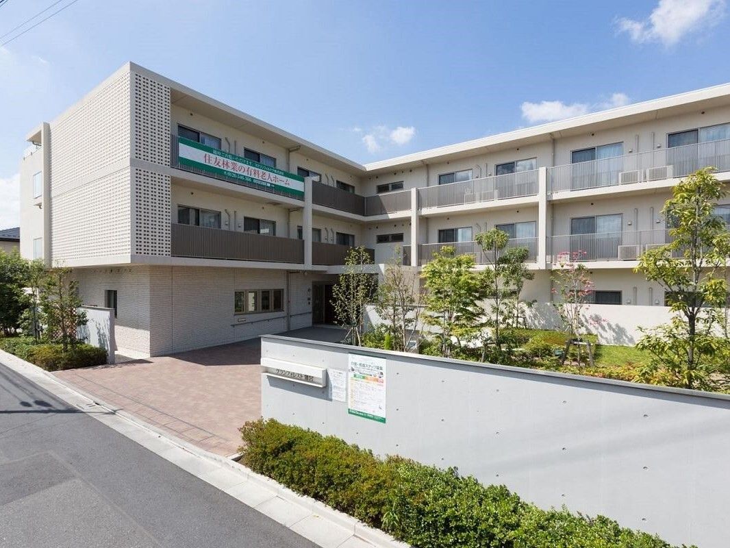 全体的にベージュ系の色でまとめられたデザインの外観である。周辺には緑の木々が植えられている。駐車場に白い車がある。