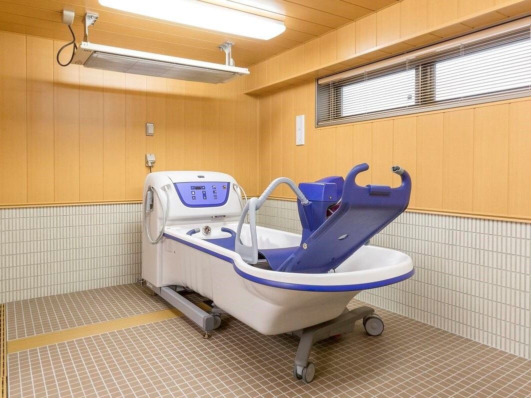 広々としたスペースが確保されている浴室には、淡いピンク色の浴室用の車イスが置いてある。浴槽は木目調デザインである。
