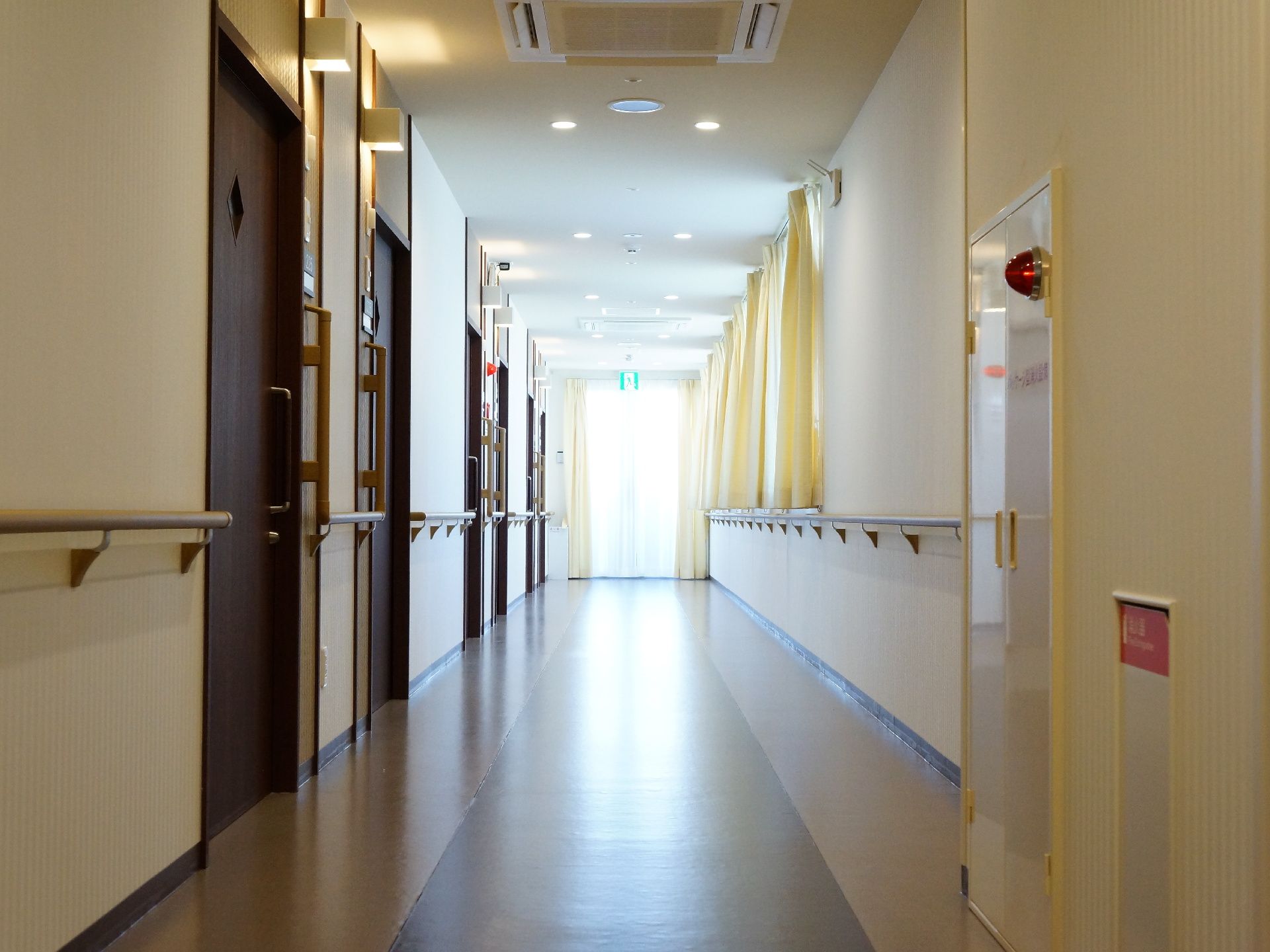 非常口から明るく光が差し込む広々とした廊下。片側は窓に面し、窓にはクリーム色のカーテンが取り付けられている。