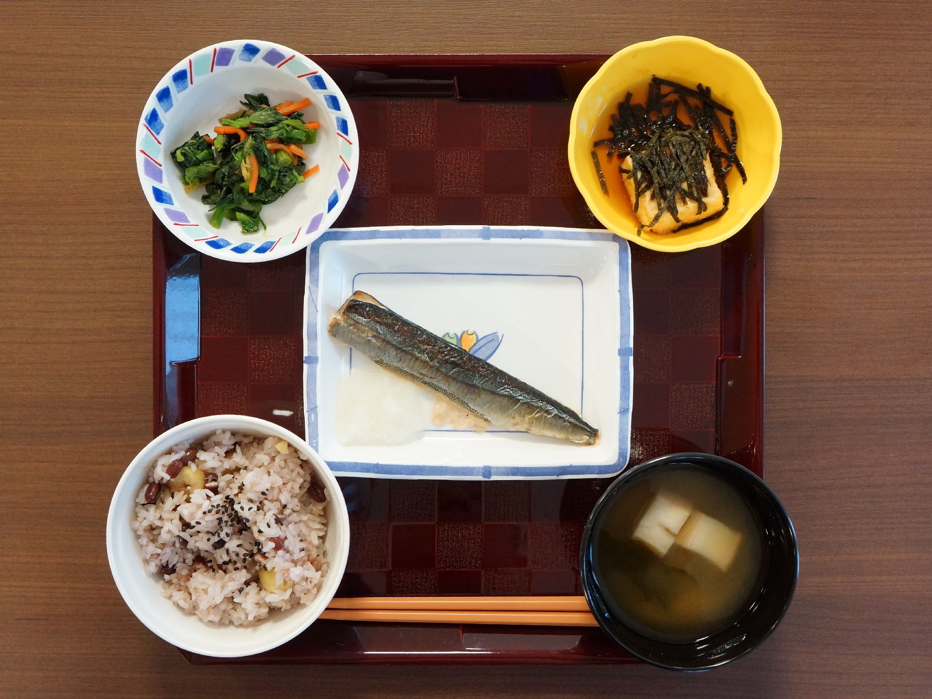 茶色いお盆の上にはお箸のほか、ごはん、お味噌汁、おかず3品が置かれている。混ぜご飯や野菜、魚で健康を考えられた献立である。
