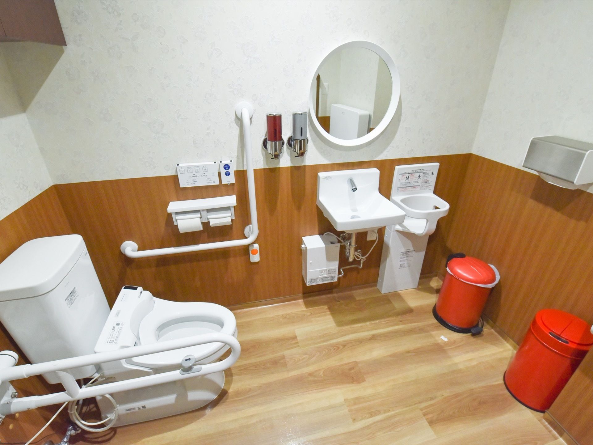 木目調の床で鏡や手洗い場を設置している。便座のわきに可動式の手すりがあり、壁にはＬ字型のものも設置されている。