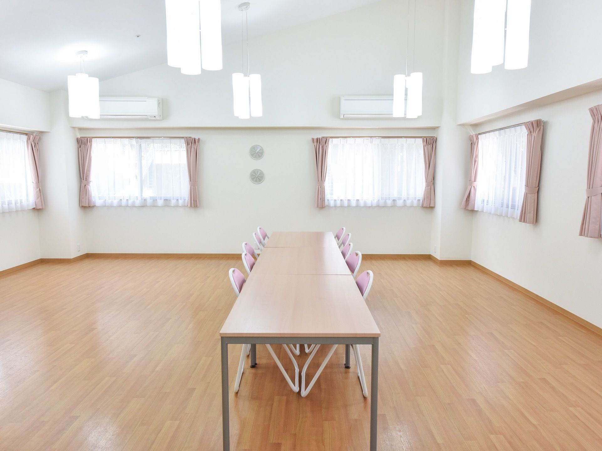 5か所以上にわたってカーテンが掛かった窓がある。広々としたフローリングの部屋の中央にはくっつけられたテーブル、椅子が置かれている。