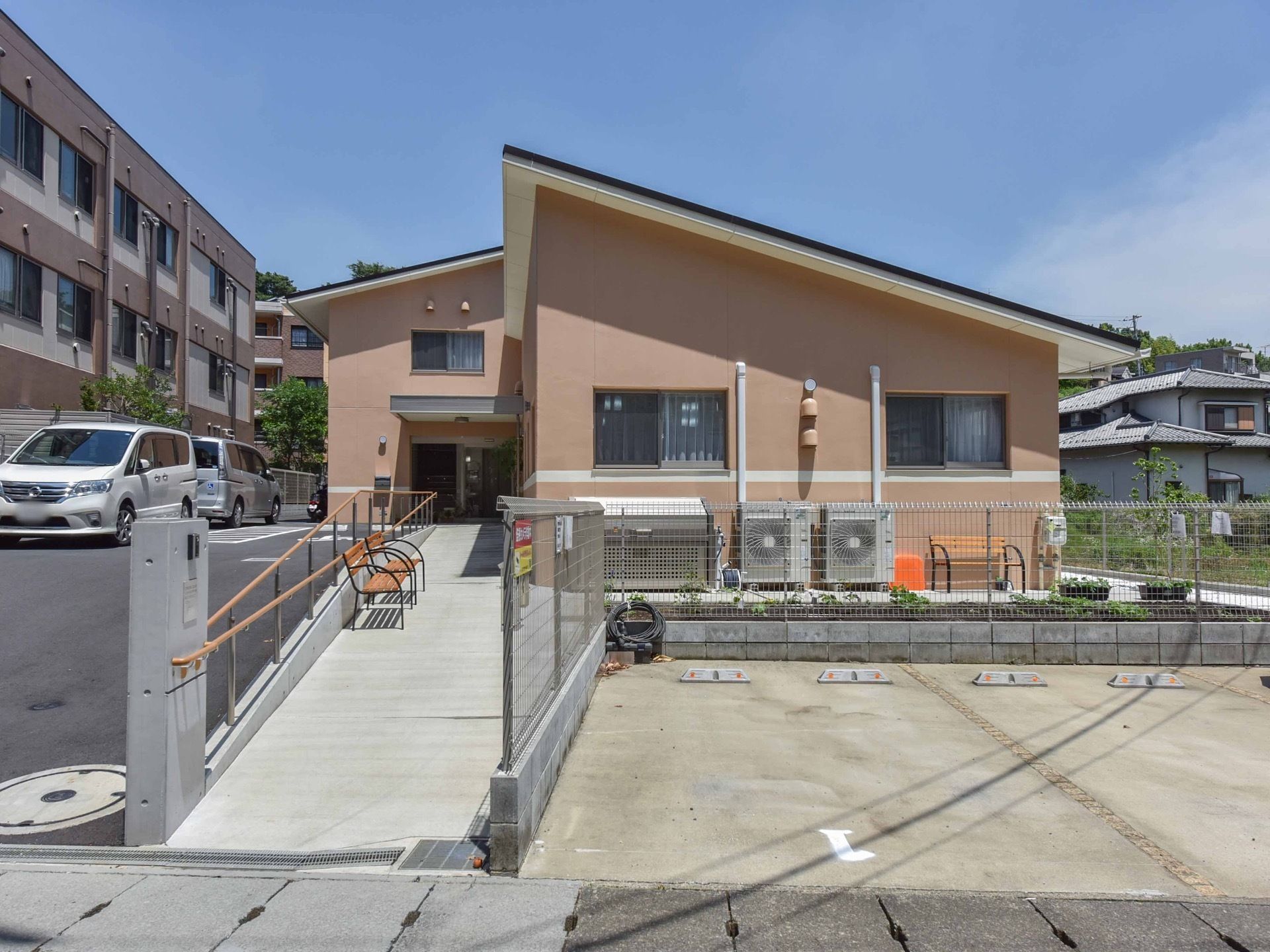 ピンクベージュ色の外壁の建物で、駐車場が整備されている。周辺道路から玄関前まで、長いスロープが設置されている。