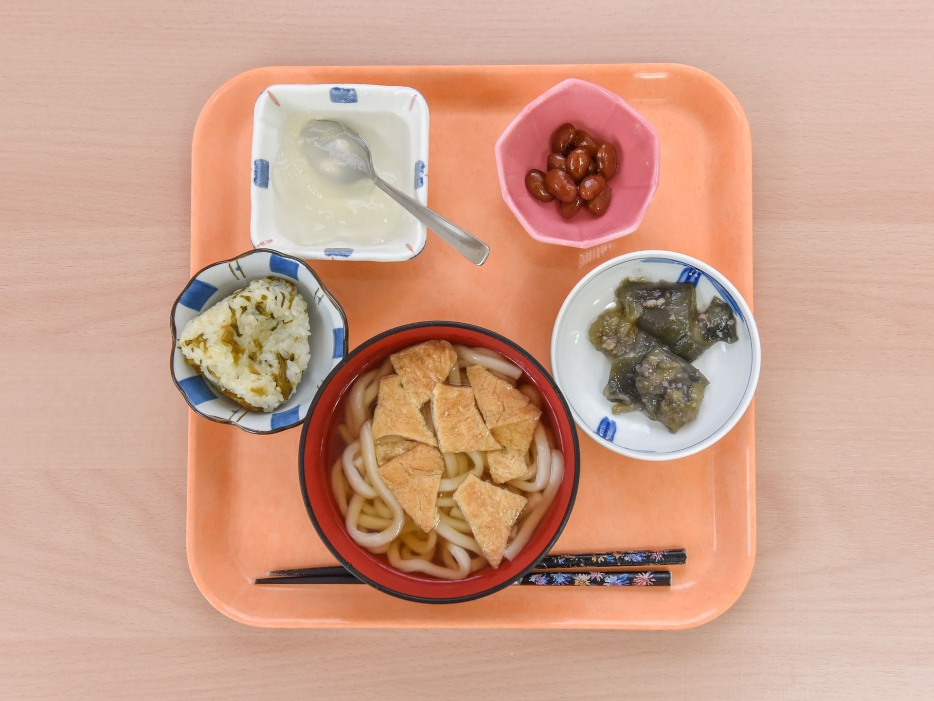 正方形のお盆の上に、食事が載っている。きつねうどんやおにぎり、豆などの副菜のほか、デザートも付いたメニューである。