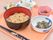 オレンジ色のトレーには、うどんの入ったお椀の脇に小鉢が３つ、煮豆やナスの漬物などが並び、四角い小鉢にデザートがスプーンを添えて置いてある。