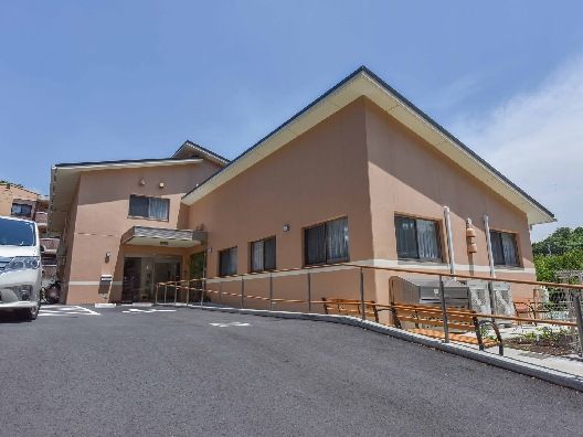 広い駐車場があり、車が止まっている。屋根が付いたブラウン系の外壁の建物の玄関にはポストがある。