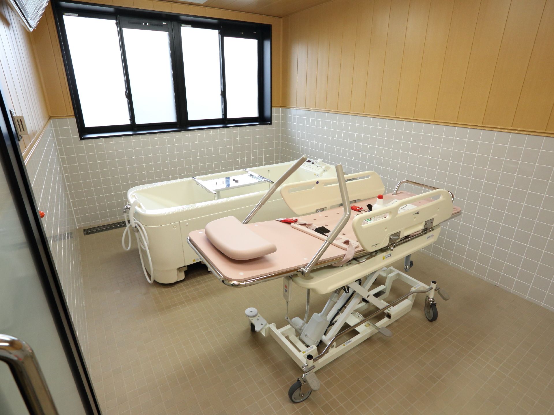 木目調とタイル調仕様の内装の浴室。奥一面は、黒縁の窓が設置されている。中央には、浴槽とストレッチャーがある。